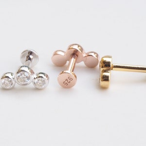 May include: Three different styles of body jewelry with a three-stone design. The first is silver with clear stones, the second is rose gold with clear stones, and the third is gold with clear stones.