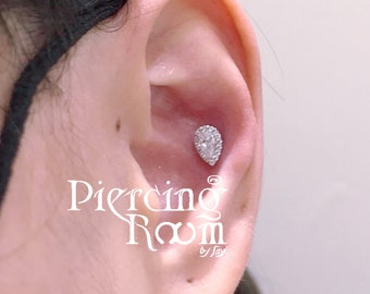 May include: A close-up of a person's ear with a small, pear-shaped diamond earring. The earring is set in a silver stud and has a halo of smaller diamonds around it. The text "Piercing Room" is visible in the lower right corner of the image.