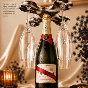 Peut inclure: Une bouteille de champagne avec un ruban rouge et une feuille d'or est présentée avec trois verres à vin suspendus à un support décoratif. Le support est noir et or. Une bougie allumée et un bol de flocons d'or sont au premier plan.