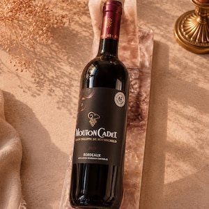 Peut inclure: Une bouteille de vin Mouton Cadet Bordeaux, avec une étiquette sombre et un bouchon rouge, repose sur un plateau effet marbre rose et blanc. La bouteille est placée devant un fond neutre avec des accents floraux séchés.