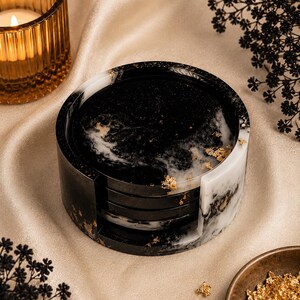 Peut inclure: Un ensemble de quatre dessous de verre noirs et blancs effet marbre avec des accents de feuilles d'or, maintenus dans un support assorti. Les dessous de verre sont ronds et reposent sur une surface en satin crème. Une bougie dans un bougeoir en verre doré est en arrière-plan.