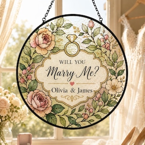 May include: A circular stained glass suncatcher featuring a floral border of pink roses and green leaves. The center displays a diamond ring icon, the text "WILL YOU Marry Me?", a small heart, and the names "Olivia & James" in black script.