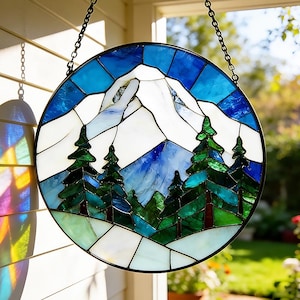 Könnte beinhalten: Runde Glasmalerei mit einer Berglandschaft mit schneebedeckten Gipfeln, immergrünen Bäumen und blauem Himmel. Das Kunstwerk ist schwarz gerahmt und hängt an einer Kette. Das Glas verwendet Blau-, Grün- und Weißtöne.