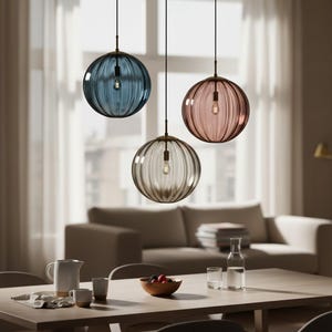May include: Three pendant lights with ribbed glass shades in blue, pink, and clear. Each globe-shaped light hangs from a black cord and features a brass-coloured fixture. The lights are suspended above a wooden dining table.