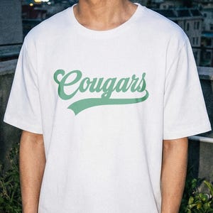 Peut inclure: T-shirt blanc avec le mot "Cougars" en vert en travers de la poitrine. Le texte a un léger effet d'ombre. Le t-shirt a des manches courtes et un col rond.