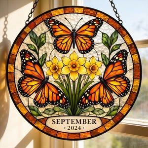 May include: A round stained glass window featuring three orange and black butterflies and yellow daffodils. The design includes green leaves and the text "SEPTEMBER 2024". The window is framed in brown and hangs from a chain.