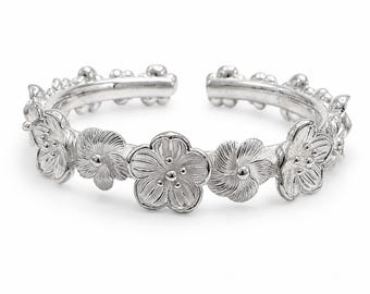 May include: A silver-toned open ring featuring a continuous band of sculpted five-petaled flowers. The metal surface has a polished finish with detailed textures on each petal and small spherical accents along the inner edge of the band.