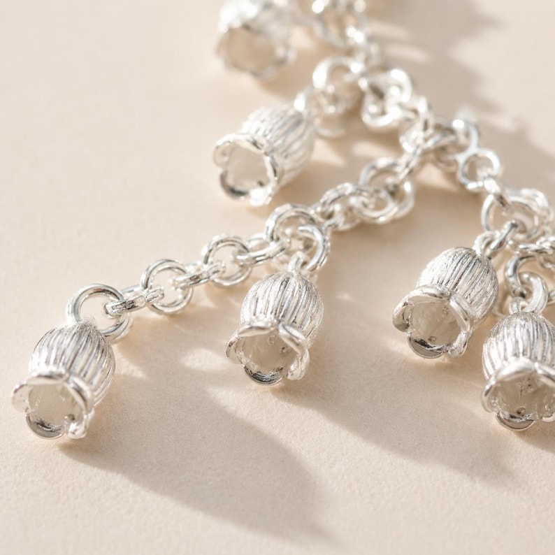 May include: A silver-toned metal chain necklace featuring multiple dangling bell-shaped flower charms. Each charm has a textured, ribbed surface and a flared opening, suspended from a series of interconnected circular links in a bright metallic finish.