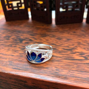 Puede incluir: Un anillo de plata con un diseño de flor de loto azul. El anillo presenta una banda calada de filigrana y una vibrante flor de loto de esmalte azul en el centro. El anillo se exhibe sobre una superficie de madera.
