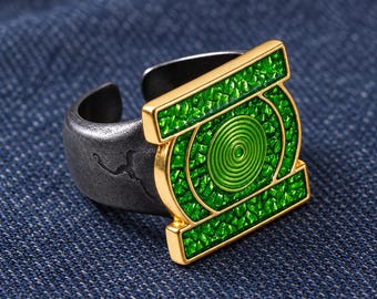 Può includere: Un anello con una fascia in metallo grigio scuro testurizzato e apertura sul retro. La parte frontale dell'anello è un emblema rettangolare dorato con una spirale circolare verde centrale circondata da un motivo a intarsio verde brillante.