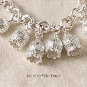 May include: A silver charm bracelet featuring multiple bell-shaped lily of the valley flower charms attached to a thick circular link chain. Each metallic flower charm displays vertical ribbed textures and scalloped edges. Text reads Lily of the Valley Details.