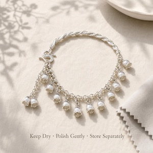 May include: A silver charm bracelet featuring a twisted chain and multiple dangling lily of the valley flower charms. The bracelet includes an S-hook clasp. Text at the bottom reads: Keep Dry &middot; Polish Gently &middot; Store Separately.