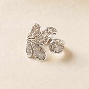 May include: A silver ring featuring an open, adjustable band design. The focal point consists of multiple teardrop-shaped elements arranged in a fan-like pattern, each filled with intricate, fine wire filigree work in a metallic silver finish.