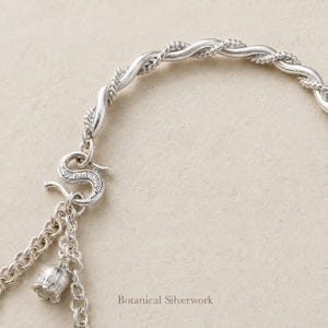 May include: A silver bracelet featuring a twisted rope design and a textured S-shaped hook clasp. A small bell-shaped charm hangs from a link chain attached to the clasp. The text Botanical Silverwork is printed in the lower center of the image.
