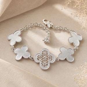 May include: A silver-toned bracelet featuring four solid, textured clover-shaped charms and one central openwork clover charm with a geometric pattern. The bracelet includes a round link chain, a small dangling charm, and a hook-style clasp closure.
