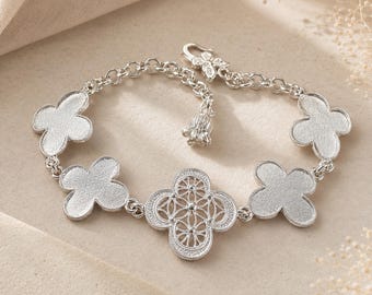 May include: A silver-toned bracelet featuring four solid, textured clover-shaped charms and one central openwork clover charm with a geometric pattern. The bracelet includes a round link chain, a small dangling charm, and a hook-style clasp closure.