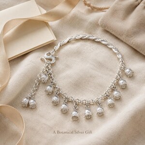May include: A silver charm bracelet featuring multiple bell-shaped flower pendants hanging from a twisted chain. The bracelet includes an S-shaped clasp and is accompanied by the text A Botanical Silver Gift printed at the bottom of the image.