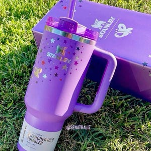 May include: A purple Stanley Quencher H2.0 tumbler with a handle, straw, and lid, decorated with gold stars and butterflies. The tumbler is next to a matching purple box with the Stanley logo. The tumbler is on a bed of green grass.