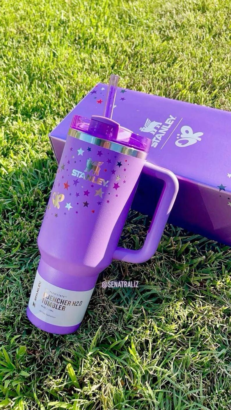May include: A purple Stanley Quencher H2.0 tumbler with a handle, straw, and lid, decorated with gold stars and butterflies. The tumbler is next to a matching purple box with the Stanley logo. The tumbler is on a bed of green grass.