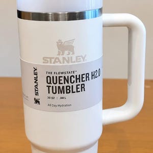 May include: A white Stanley Quencher H2.0 tumbler with a handle, a clear straw, and a lid. The product label reads: Stanley The Flowstate Quencher H2.0 Tumbler 30 oz | .88 L All Day Hydration. The tumbler features a metallic band below the lid.