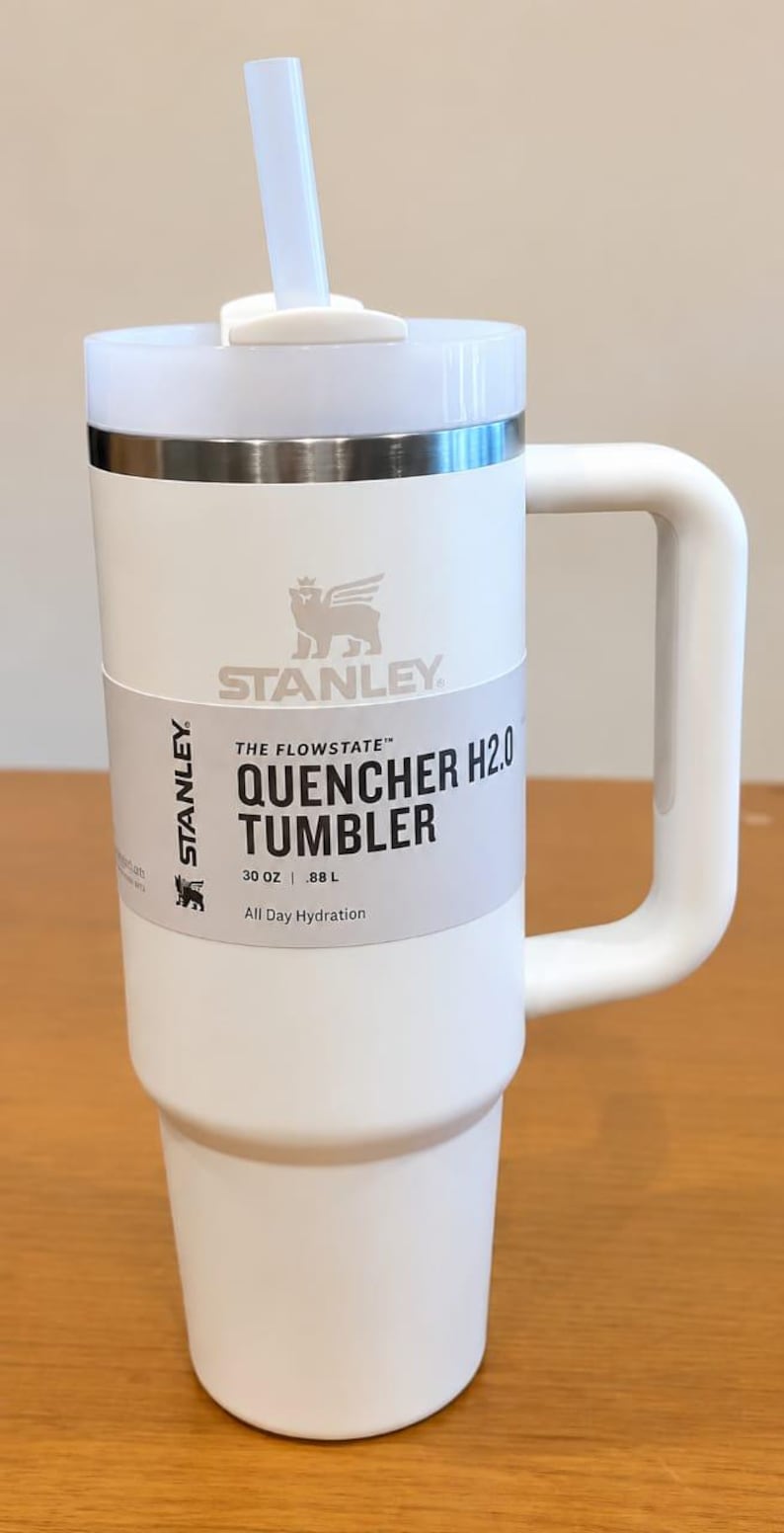 May include: A white Stanley Quencher H2.0 tumbler with a handle, a clear straw, and a lid. The product label reads: Stanley The Flowstate Quencher H2.0 Tumbler 30 oz | .88 L All Day Hydration. The tumbler features a metallic band below the lid.