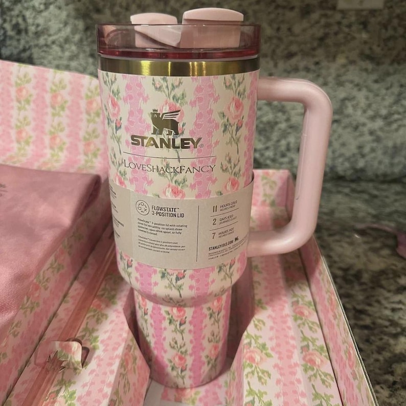 K&ouml;nnte beinhalten: Ein pink-wei&szlig;es Stanley-Trinkgef&auml;&szlig; mit floralem Muster, Griff und Deckel. Das Trinkgef&auml;&szlig; hat die Logos von Stanley und Loveshackfancy. Die Verpackung ist ebenfalls pink und wei&szlig; floral.