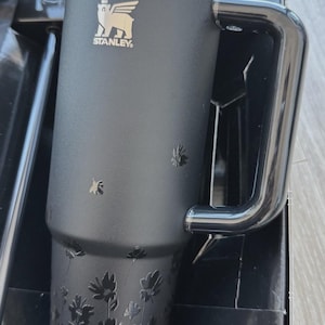 May include: Black Stanley travel mug with a handle and a floral design. The mug features the Stanley logo and a black lid. The mug is in its original packaging. The mug is designed to hold approximately 20 ounces.