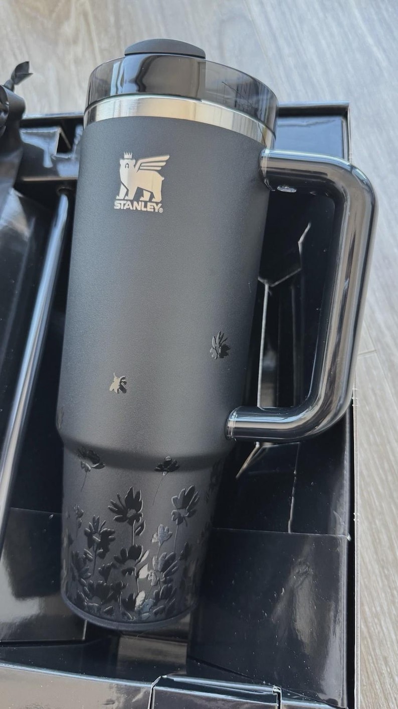 May include: Black Stanley travel mug with a handle and a floral design. The mug features the Stanley logo and a black lid. The mug is in its original packaging. The mug is designed to hold approximately 20 ounces.