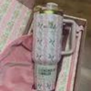 K&ouml;nnte beinhalten: Ein rosa-wei&szlig;er isolierter Becher mit Griff und goldfarbenem Deckel. Der Becher hat ein Blumenmuster und den Text "Forever by Stanley". Er wird in einer rosa Geschenkbox mit passendem rosa Stofffutter pr&auml;sentiert.