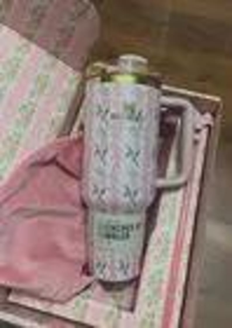 K&ouml;nnte beinhalten: Ein rosa-wei&szlig;er isolierter Becher mit Griff und goldfarbenem Deckel. Der Becher hat ein Blumenmuster und den Text "Forever by Stanley". Er wird in einer rosa Geschenkbox mit passendem rosa Stofffutter pr&auml;sentiert.