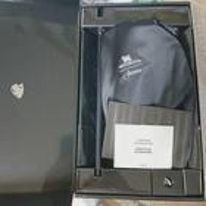May include: An open black box with a black stand, a black bag with a logo, and a white card. The box has a silver heart-shaped emblem on the upper left. The bag has white text. The box is on a light gray surface.