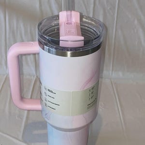 K&ouml;nnte beinhalten: Ein hoher, zylindrischer Becher mit hellrosa-wei&szlig;em Marmormuster, ausgestattet mit einem gro&szlig;en rosa Seitengriff, einem durchsichtigen Kunststoffdeckel mit rosa Drehverschluss und einem transparenten Strohhalm. Ein Etikett umschlie&szlig;t die Mitte.