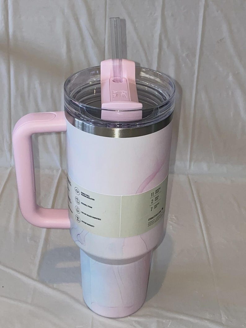 K&ouml;nnte beinhalten: Ein hoher, zylindrischer Becher mit hellrosa-wei&szlig;em Marmormuster, ausgestattet mit einem gro&szlig;en rosa Seitengriff, einem durchsichtigen Kunststoffdeckel mit rosa Drehverschluss und einem transparenten Strohhalm. Ein Etikett umschlie&szlig;t die Mitte.