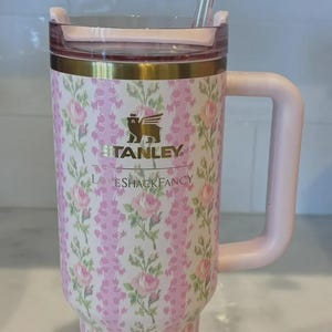 K&ouml;nnte beinhalten: Ein Stanley-Becher mit rosa und wei&szlig;en Blumen, passendem Griff und Strohhalm. Der Becher tr&auml;gt die Aufschrift "STANLEY" und "LOVESHACKFANCY". Der Strohhalm hat eine dekorative Schleife.