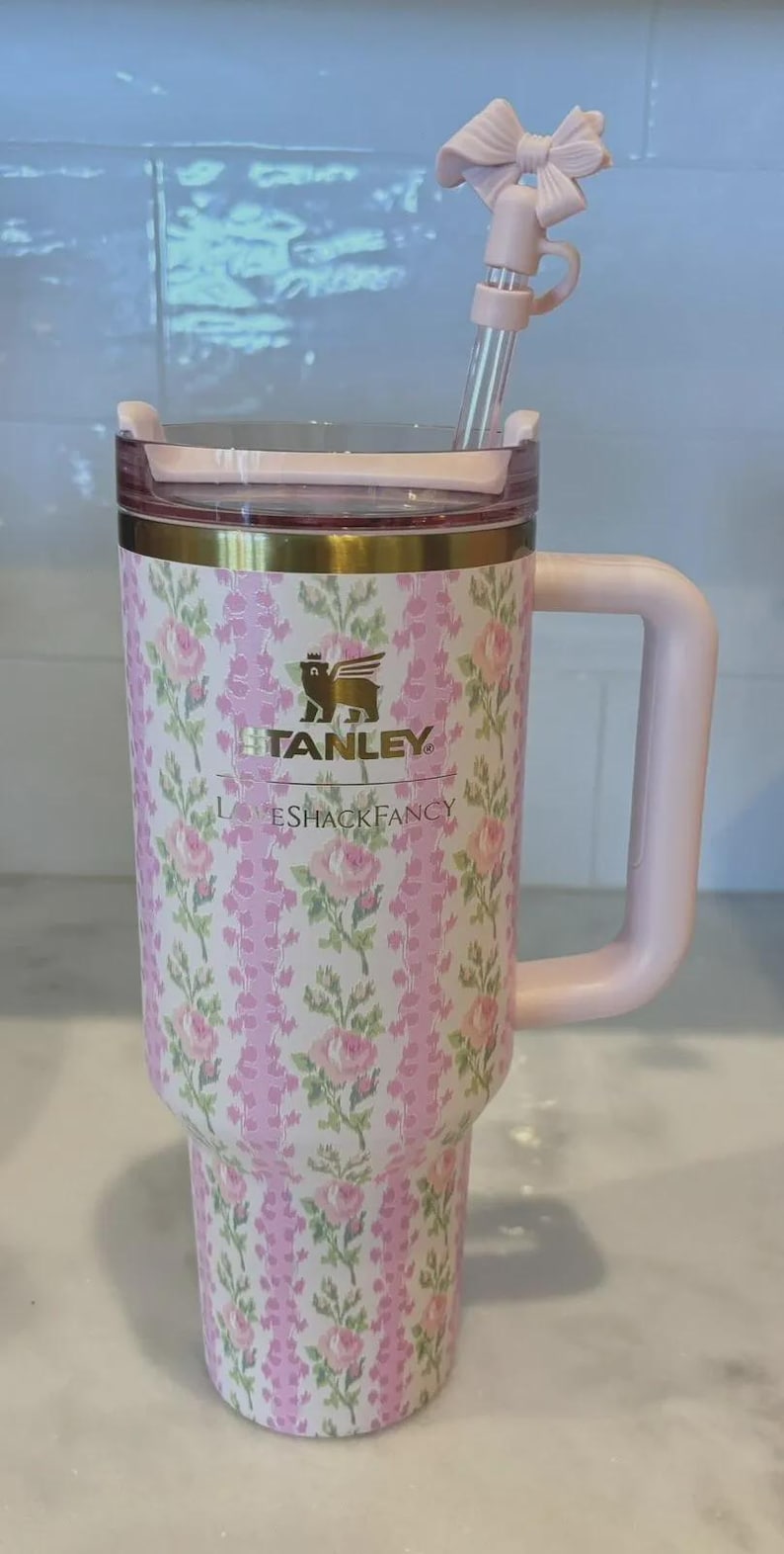 K&ouml;nnte beinhalten: Ein Stanley-Becher mit rosa und wei&szlig;en Blumen, passendem Griff und Strohhalm. Der Becher tr&auml;gt die Aufschrift "STANLEY" und "LOVESHACKFANCY". Der Strohhalm hat eine dekorative Schleife.