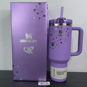 May include: A purple Stanley Quencher H2.0 FlowState tumbler with a handle and straw, next to a matching box. The tumbler and box are decorated with star and butterfly graphics. The Stanley logo is visible on both items.
