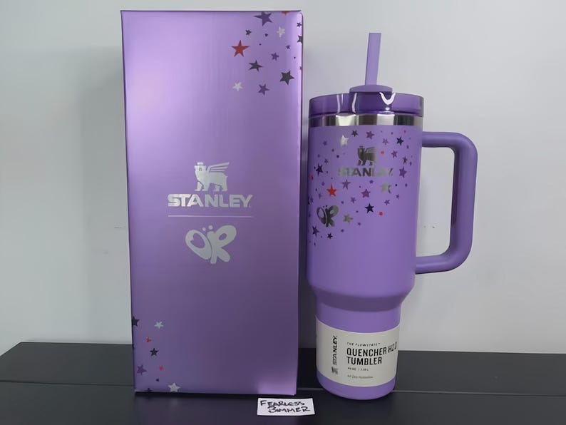 May include: A purple Stanley Quencher H2.0 FlowState tumbler with a handle and straw, next to a matching box. The tumbler and box are decorated with star and butterfly graphics. The Stanley logo is visible on both items.