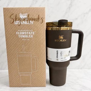 Puede incluir: Un vaso t&eacute;rmico marr&oacute;n Stanley Quencher H2.0 Flowstate con una capacidad de 1,18 L y asa a juego, junto a su caja de cart&oacute;n. La caja muestra el texto Skyhawks, Stanley y The Quencher H2.0 Flowstate Tumbler 40 oz 1,18 L.