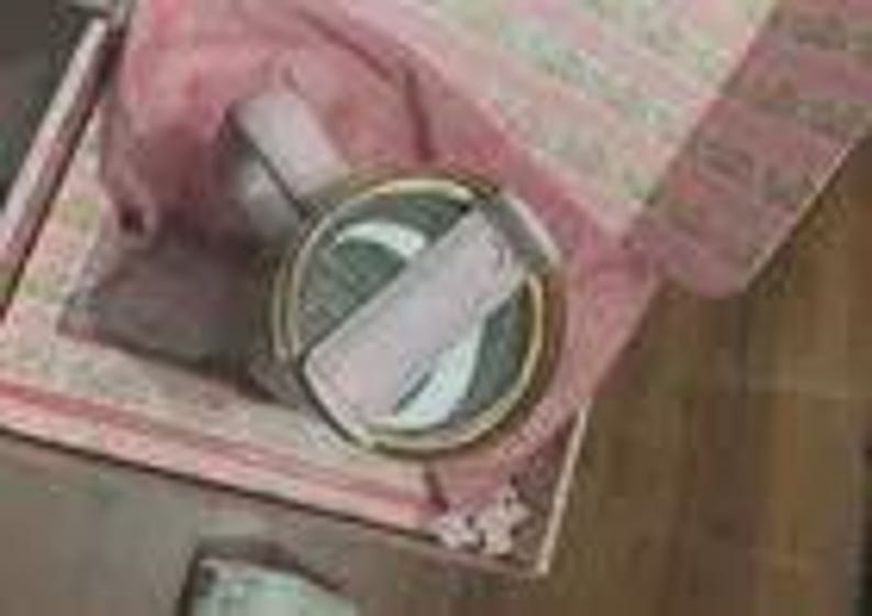 K&ouml;nnte beinhalten: Eine dekorative, runde Schmuckschatulle mit goldenem Rand und hellrosa Deckel. Die Schatulle ist ge&ouml;ffnet und zeigt einen hellrosa Schal und einen kleinen, runden Gegenstand im Inneren. Die Schatulle befindet sich in einer gr&ouml;&szlig;eren, gemusterten Schachtel.