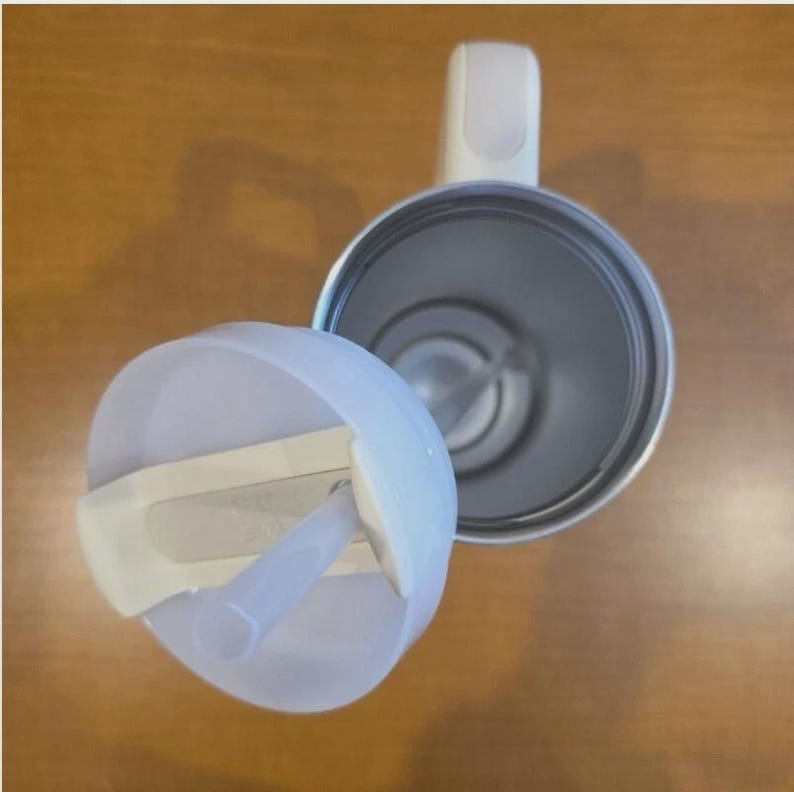 May include: A top-down view of a white tumbler with a matching lid and a clear straw. The tumbler features a white handle attached to the side, and the interior is metallic silver. The lid is partially detached, revealing the open circular top.