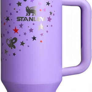May include: A lavender Stanley Quencher tumbler with a handle and straw. The tumbler is decorated with star and butterfly graphics. The Stanley logo is visible on the front. The tumbler is designed to keep drinks cold for hours.