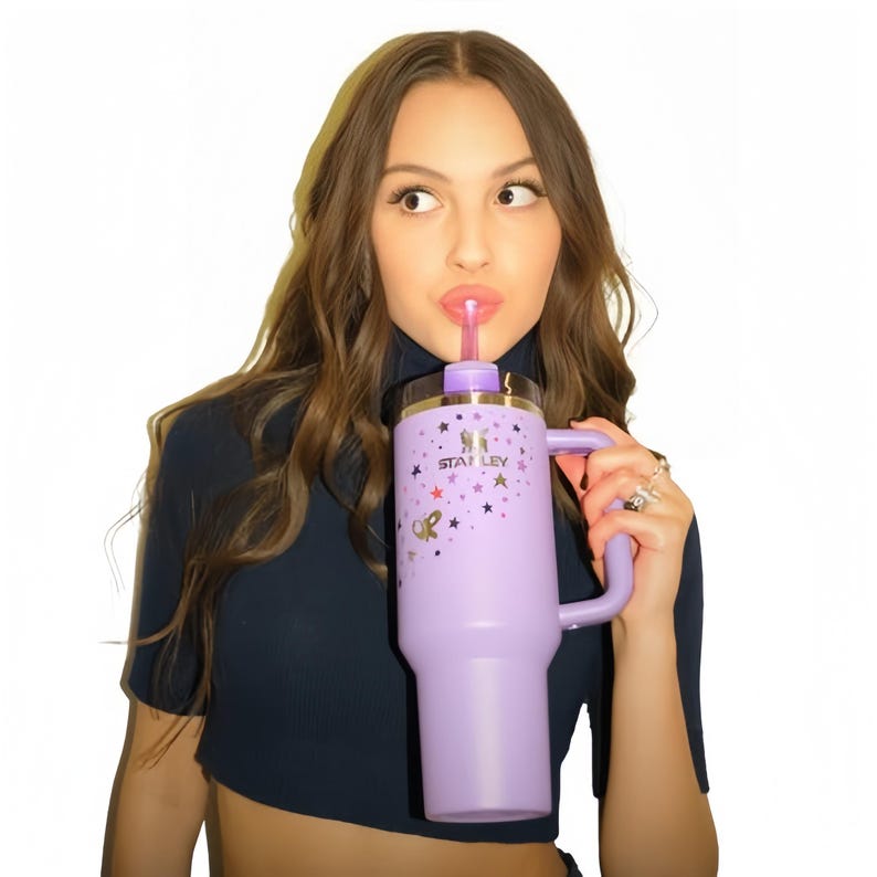 May include: A lavender Stanley tumbler with a handle, a straw, and a gold rim. The tumbler is decorated with gold stars and the Stanley logo. The person is wearing a dark blue top.