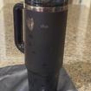 May include: A black insulated tumbler with a handle and a clear lid. The tumbler has a stainless steel rim and a logo on the side. It is sitting on a black cloth.