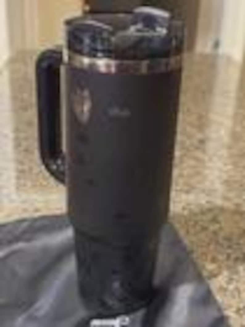 May include: A black insulated tumbler with a handle and a clear lid. The tumbler has a stainless steel rim and a logo on the side. It is sitting on a black cloth.