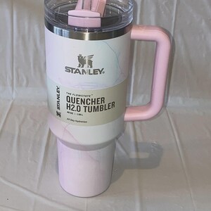 K&ouml;nnte beinhalten: Ein 1,18 L Stanley Quencher H2.0 Becher mit hellrosa-wei&szlig;em Marmormuster. Der Becher verf&uuml;gt &uuml;ber einen rosa Griff, einen transparenten Deckel mit rosa Drehverschluss und einen durchsichtigen Strohhalm. Ein Produktetikett ist mittig angebracht.
