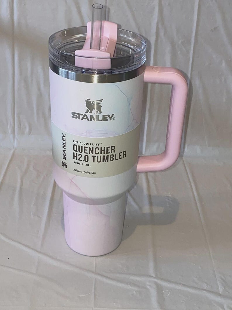 K&ouml;nnte beinhalten: Ein 1,18 L Stanley Quencher H2.0 Becher mit hellrosa-wei&szlig;em Marmormuster. Der Becher verf&uuml;gt &uuml;ber einen rosa Griff, einen transparenten Deckel mit rosa Drehverschluss und einen durchsichtigen Strohhalm. Ein Produktetikett ist mittig angebracht.