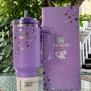 May include: A purple Stanley Flowstate Quencher H2.0 tumbler with a handle and straw, and a matching box. The tumbler and box are decorated with silver and colorful star designs. The tumbler has a 40 oz capacity.