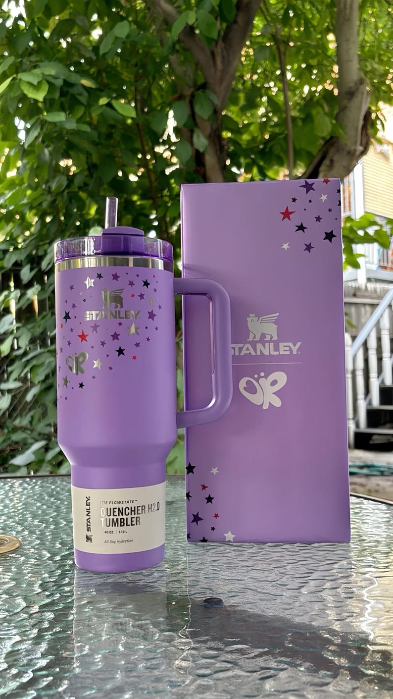 May include: A purple Stanley Flowstate Quencher H2.0 tumbler with a handle and straw, and a matching box. The tumbler and box are decorated with silver and colorful star designs. The tumbler has a 40 oz capacity.
