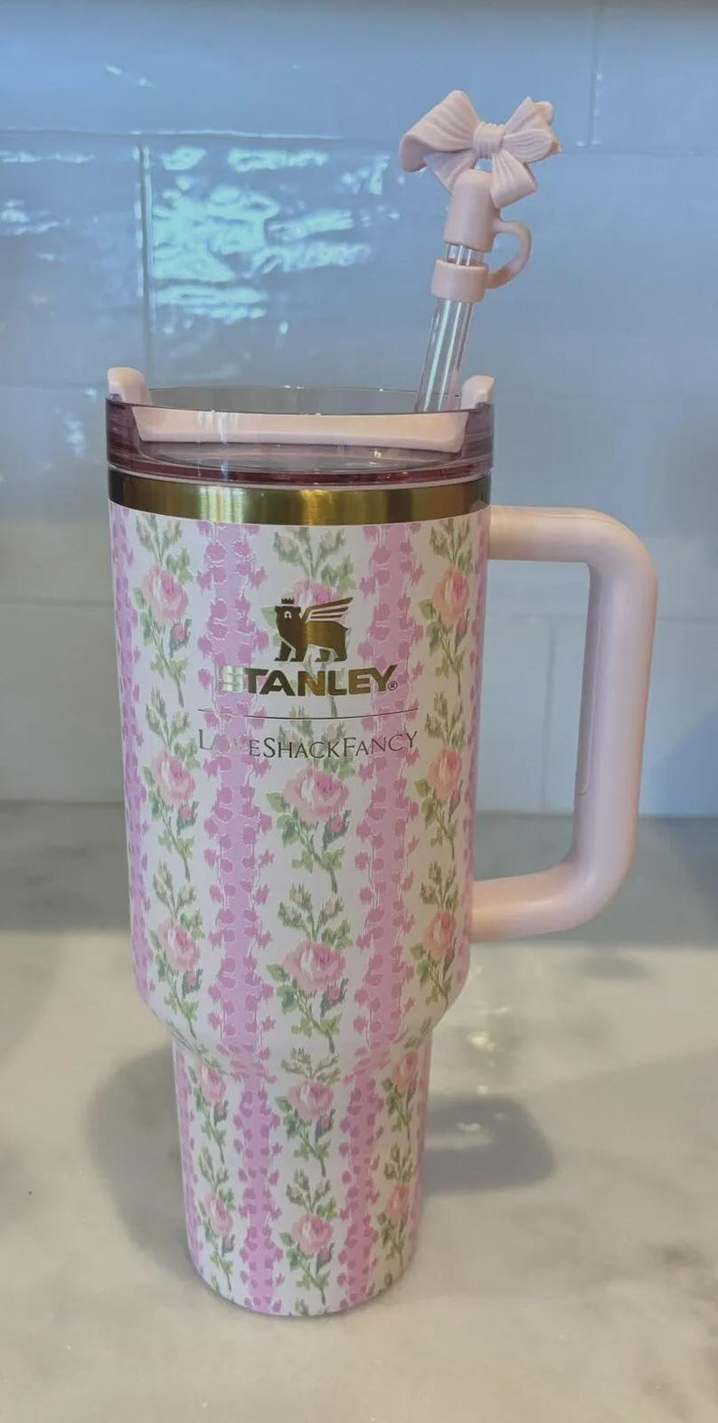 K&ouml;nnte beinhalten: Ein Stanley-Becher mit rosa und wei&szlig;en Blumen, passendem Griff und Strohhalm. Der Becher tr&auml;gt die Aufschrift "STANLEY" und "LOVESHACKFANCY". Der Strohhalm hat eine dekorative Schleife.