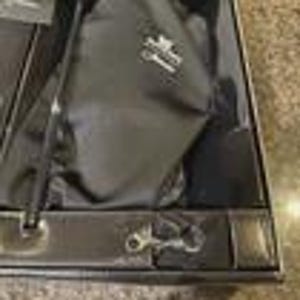 May include: A black golf glove and accessories are displayed inside a black box. The glove has a silver emblem. A black tee and a silver-colored key ring are also visible. The box is open, revealing the contents.